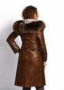 Red-haired model posing with her back to us in luxury brown coat of crocodile skin with fur hood and wide leather belt