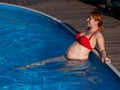 A red-haired pregnant woman in a red bikini is resting in the pool, leaning on the side. Royalty Free Stock Photo