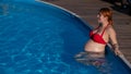 A red-haired pregnant woman in a red bikini is resting in the pool, leaning on the side. Royalty Free Stock Photo