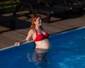 A red-haired pregnant woman in a red bikini is resting in the pool, leaning on the side. Royalty Free Stock Photo