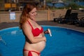 A red-haired pregnant woman sunbathes by the pool. The expectant mother applies sunscreen to her tummy. Royalty Free Stock Photo