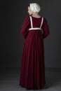 A Regency maid servant wearing a red linen dress with an apron and cap against a studio backdrop