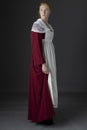 A Regency maid servant wearing a red linen dress with an apron and a lace modesty shawl against a studio backdrop