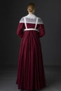 A Regency maid servant wearing a red linen dress with an apron and a lace modesty shawl against a studio backdrop