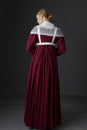 A Regency maid servant wearing a red linen dress with an apron and a lace modesty shawl against a studio backdrop