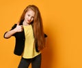 Red-haired schoolgirl in black jacket and pants, yellow t-shirt. She is showing thumb up, posing on orange background. Close up