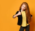 Red-haired schoolgirl in black jacket and pants, yellow t-shirt. She is showing thumb up, posing on orange background. Close up