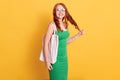 Red haired smiling girl with white blank cotton bag, looks at camera and pulling her curl, wearing green dress, standing against Royalty Free Stock Photo