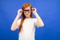 Red-haired teenager girl squints while holding glasses in her hand on a blue studio background