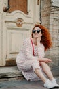 Red haired thoughtful woman has rest after strolling, poses near old building, sits on threshold, wears sunglasses, dress and