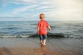Toddler child kid in red t-shirt and jeans standing near water on lake sea ocean beach at evening sunset looking far away. Royalty Free Stock Photo