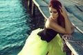 Red haired woman in black corset and long tail green veiling skirt standing in the sea water leaning on the pier. Mermaid concept