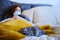 A red haired woman in a medical mask sick with the flu virus and a cat lie on the bed Royalty Free Stock Photo