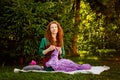 A red-haired woman is sitting on a blanket in the park and crocheting a lilac shawl. Hobby classes in nature Royalty Free Stock Photo