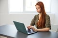 red-haired woman typing in a laptop sitting at a table in the office near the window working full time Royalty Free Stock Photo