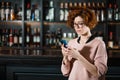 Red-haired woman using smartphone in restaurant standing near bar stand Royalty Free Stock Photo