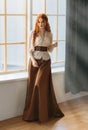 Red-haired woman in vintage dress stands at large classic window waiting love. Clothing costume countess old style white Royalty Free Stock Photo