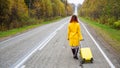 A red-haired woman in a yellow coat walks with a yellow suitcase along the highway in autumn. Royalty Free Stock Photo