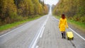 A red-haired woman in a yellow coat walks with a yellow suitcase along the highway in autumn. Royalty Free Stock Photo
