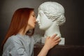 Red-haired young girl on a black background a gray t-shirt, kissing with a plaster bust Royalty Free Stock Photo