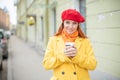 The red-haired young woman in a yellow coat and red beret is drinking coffee on the street to keep warm Royalty Free Stock Photo