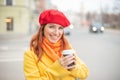 The red-haired young woman in a yellow coat and red beret is drinking coffee on the street to keep warm Royalty Free Stock Photo