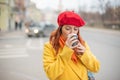 The red-haired young woman in a yellow coat and red beret is drinking coffee on the street to keep warm Royalty Free Stock Photo