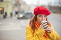 The red-haired young woman in a yellow coat and red beret is drinking coffee on the street to keep warm Royalty Free Stock Photo