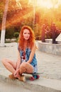 Red haired young women sitting on skateboard with her legs crossed backlit by sun