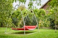 Red hammock swing in metal frame with nobody on green lawn in backyard near log house cottage. Rest relax relaxation alone on