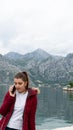 Red head young girl talking by cell phone with red jacket in a lake. Kotor bay with grey mountains in a lake