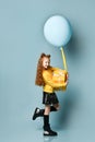 Teen child in yellow sweatshirt, black skirt, knee-highs, boots. Smiling, holding balloon and gift box, posing on blue background.