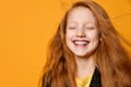 Red-headed teenage child in black jacket and yellow t-shirt. She laughing with closed eyes, posing on orange background. Close up