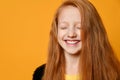 Red-headed teenage child in black jacket and yellow t-shirt. She laughing with closed eyes, posing on orange background. Close up