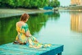 Red-headed woman in bright summer dress with open back sitting on the wooden pier at the river bank