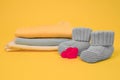 Red hearts, knitted booties for newborn baby and heap on clothes on yellow background. Expecting a baby concept. Selective focus.