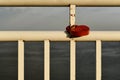 A red metallic lock in the shape of a heart hangs on a rusty white lattice of a river embankment railing.