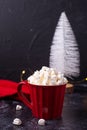 Red mug with hot cacao and heap of mashmellows with white decorative tree on black textured background.