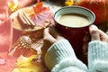 Red mug with hot cocoa on table with fallen maple leaves, round lamps, garlands, cinnamon sticks, warm blanket. Autumn atmosphere,