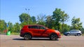 A red new car is driving along the road, standing out in the general traffic. A park with green trees, road signs, and a Royalty Free Stock Photo