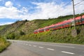 A red passenger train driving alongside an empty motorway next to the hills of a vineyard in Germany. Royalty Free Stock Photo