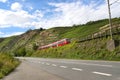 A red passenger train driving alongside an empty motorway next to the hills of a vineyard in Germany.