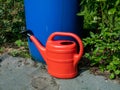 Red, plastic watering on ground can in front of blue, plastic water barrel reused for storing water for watering plants in bright