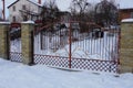 Red metal gates of forged rods with a pattern and part of a brick fence on the street in white snow Royalty Free Stock Photo