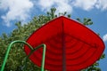 Red umbrella shaped visor with green handrail on the kid`s playground against a background of tree foliage and blue sky