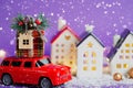 Red retro car on snow carries gift box with home key with keychain cottage on roof past houses with fairy lights, Christmas tree. Royalty Free Stock Photo