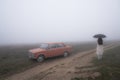 Red retro car stands near the dirt road in fog, young girl in white coat stands under umbrella Royalty Free Stock Photo