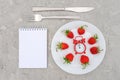 Red ripe strawberries berry on white plate, cutlery, red alarm clock and blank notepad on gray stone background table. Top view, Royalty Free Stock Photo