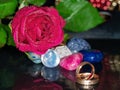 Red rose covered in water beads colorful gem stone in focus, Wedding rings in foreground out of focus. Concept marriage, romance