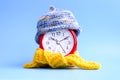 Red round alarm clock in knitted wool blue hat and yellow scarf on a blue background. winter time concept. winter season Royalty Free Stock Photo